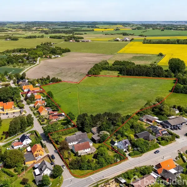 Landsby med røde hustage ligger langs en vej, omgivet af marker i forskellige nuancer af grønt og gult. En rød linje markerer et større område, der inkluderer flere bygninger.