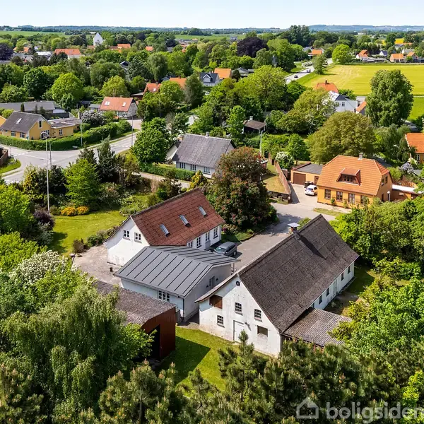 To hvide huse med røde tage står blandt grønne træer og marker i en landsby. Flere huse og veje omgiver ejendommene, mens landskabet strækker sig ud i det fjerne.