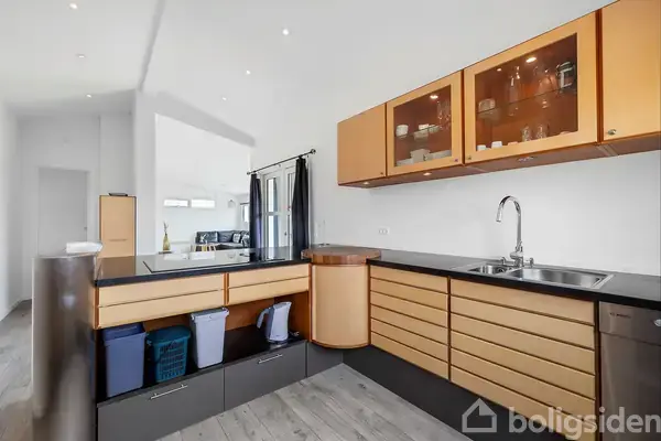 Køkken med træskabsfronter og sort bordplade inkluderer en vask med vandhane. Skuffer og skraldespande i hjørnet; glaslåger øverst. Åben forbindelse til stueområde.