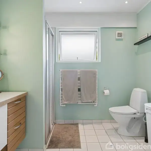 Badeværelse med hvide fliser og lysegrønne vægge. En vask med skuffer og et rundt spejl til venstre. Toilet og håndklædetørrer ved siden af et vindue. Brusekabine findes også.