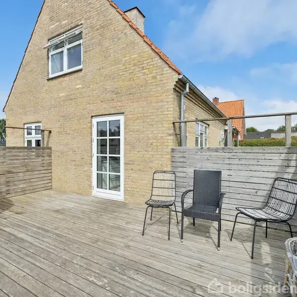 Terrasse med træbræt gulv, hvor der står tre sorte stole. En svingstol hænger til venstre. I baggrunden ses et murstenshus med en havedør og vindue.
