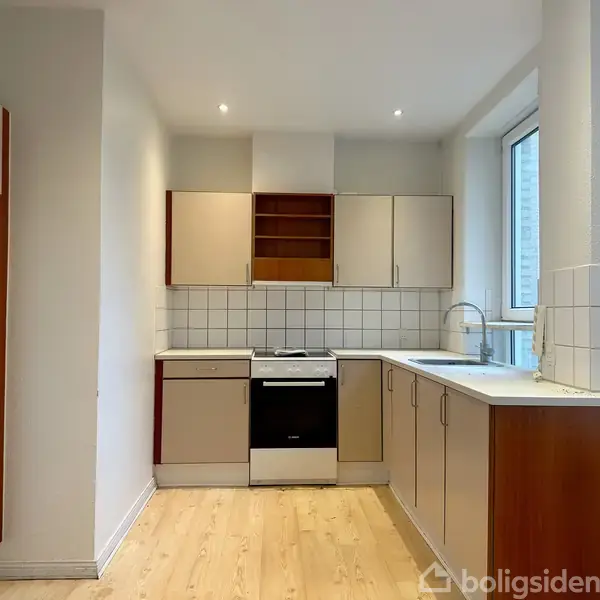 Et moderne køkken med ovn og vask, omgivet af lyse skabe og fliser, ligger i et rum med lyst trægulv og et stort vindue til venstre.