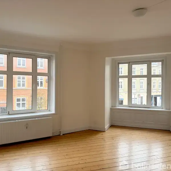 Et lyst rum med trægulv har to store vinduer, der slipper dagslys ind. Udvendigt ses facader af klassiske murstensbygninger. Væggene er hvide, og rummet er tomt.