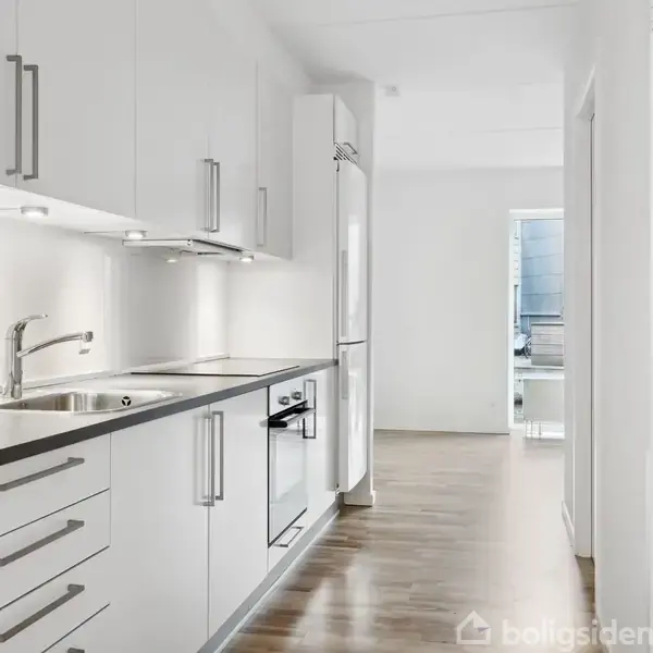 Et hvidt, moderne køkken med indbyggede skabe og apparater strækker sig langs en smal gang. Det har træparketgulv, og i baggrunden ses en lys åbning til et andet rum.