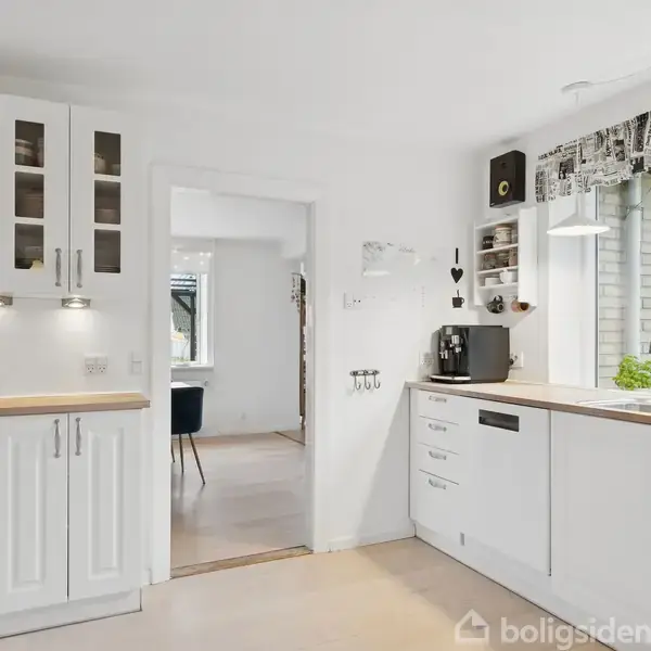 Køkken med hvide skabe og træbordplader, kaffemaskine på bord, vindue med udsigt og lysindfald. Åben dør fører til spisestue med lysindtrængning. Mørk stol ses i baggrunden.