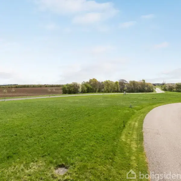 En grøn mark strækker sig langs vejen, som fører mod et hus med trimmede buske i forgrunden. I baggrunden ses træer og åbne marker under en klar himmel.