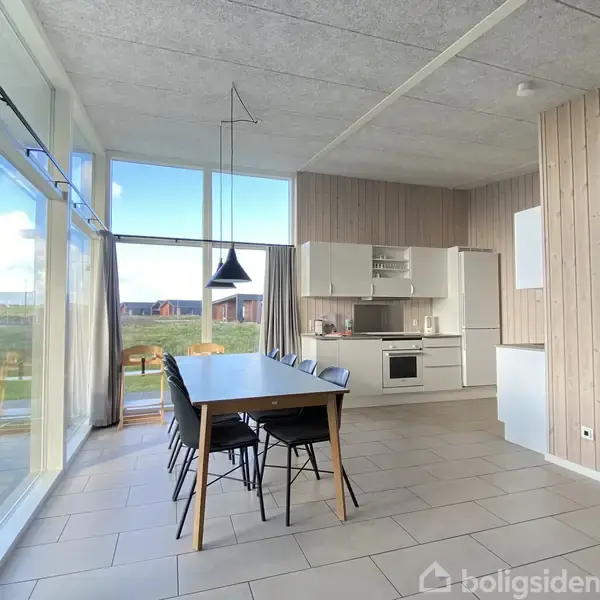 Et moderne køkken-alrum med et langt spisebord og sorte stole. Store vinduer giver udsigt til en græsplæne og flere huse i baggrunden. Enkle, hvide køkkenelementer mod en trævæg.