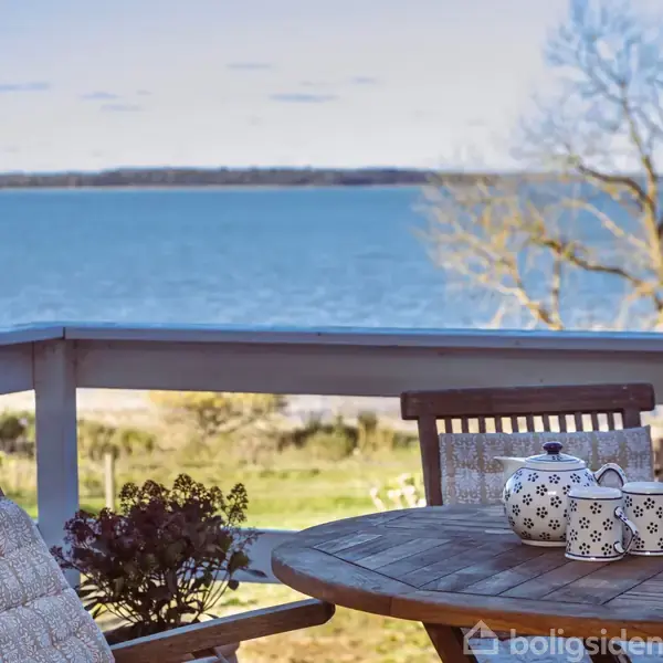 Et rundt træbord med en blomstret tekande og kop står på en terrasse med udsigt over en sø. To træstole omkranser bordet. En træeramme tilbyder panoramaudsigt.