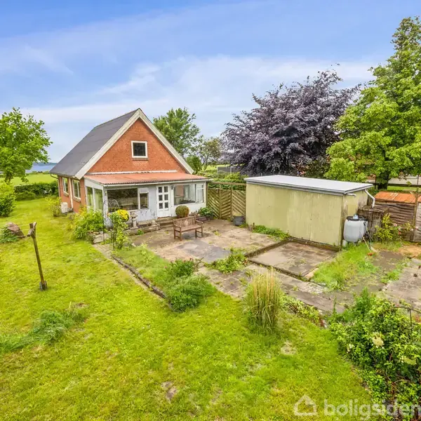 Et lille rødt hus med skråt tag står stille ved siden af en flisebelagt terrasse i en grøn have omkranset af træer og grønne marker.