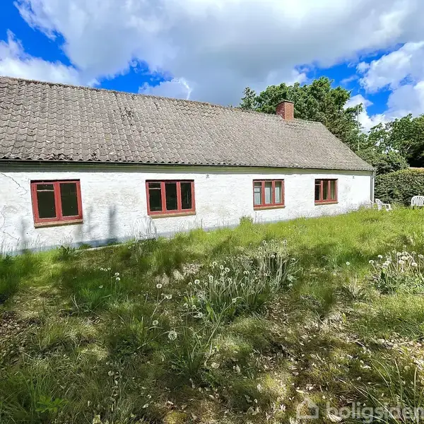 Et hvidt hus med røde vinduesrammer står i en have med højt græs. Baggrunden har grønne træer, og himlen er delvist overskyet med blå og hvide skyer.