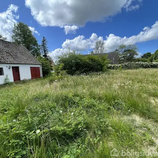 En hvid bygning med røde døre står stille i en stor, vildtvoksende græsplæne. Skyer dækker delvist en blå himmel over en frodig landskabshave.