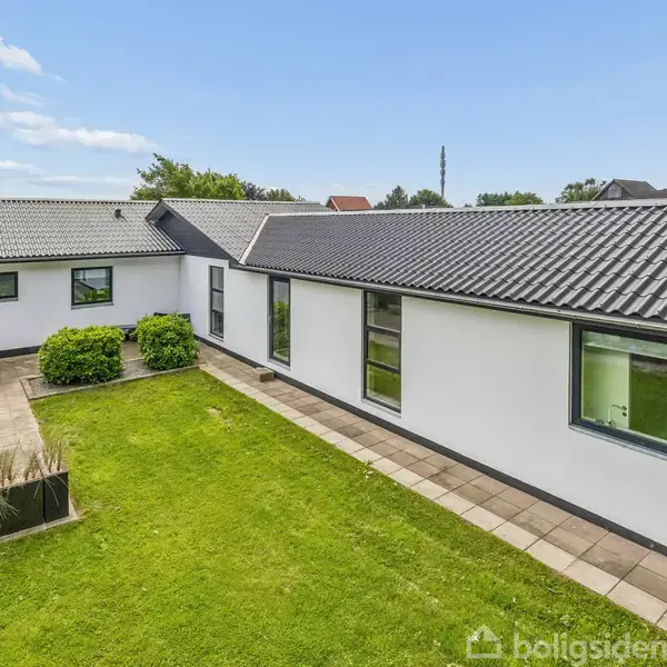 Hvidt, moderne hus med sorte tagsten, store vinduer og en L-formet struktur. Foran huset er en grøn græsplæne omgivet af fliser og en terrasse med havemøbler.