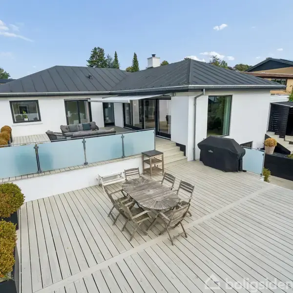 Et moderne hus med fladt tag har en stor træterrasse. Et spisebord med stole og en grill står på terrassen, omgivet af glasrækværk og plantekrukker.