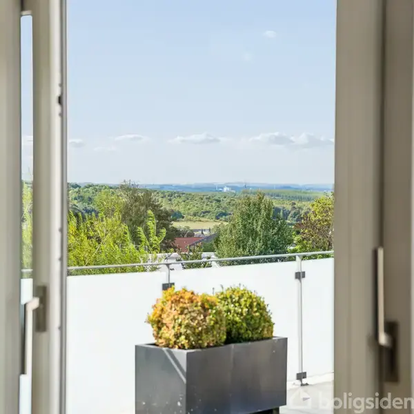 Dobbelt glasdør står åben og afslører en terrasse med planter i sorte kvadratiske plantekasser. I baggrunden ses en frodig udsigt med træer og blå himmel.