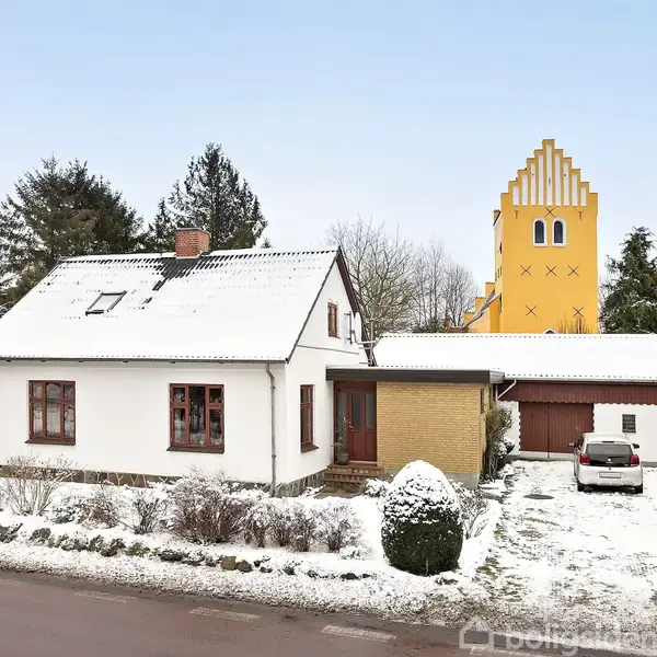 Et hvidt hus med rødt tag ligger i et snedækket område. En gul kirke med karakteristisk trappet gavl ses i baggrunden. Flere træer omgiver bygningerne.