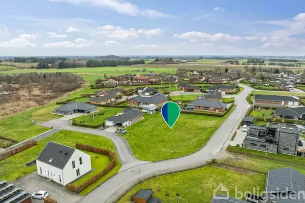 Grøn markør står over ledig byggegrund omgivet af parcelhuse. Grunden er en del af et boligområde med grønne arealer og asfalterede veje, beliggende i et landligt område.