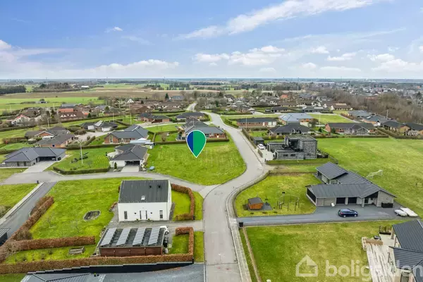 Grøn grund markeret med en nål; omgivet af moderne villaer i et boligområde. Området er frodigt med åbne landskaber i baggrunden, og vejnet forbinder husene.