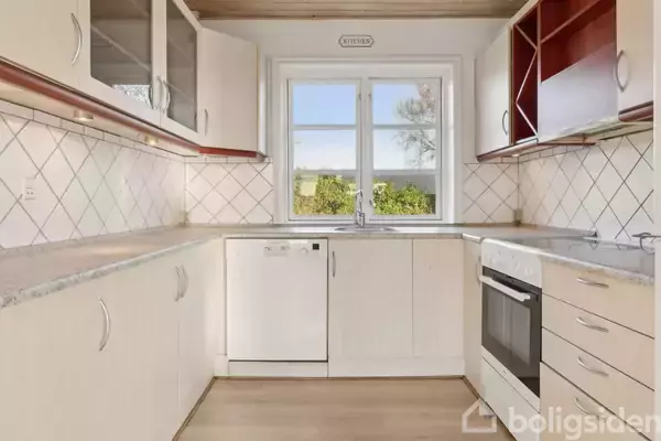 Køkken indrettet i hvidt med komfur til højre, opvaskemaskine i midten, fliser på væggen, og et vindue med udsigt til have. Skiltet over vinduet siger "KITCHEN".