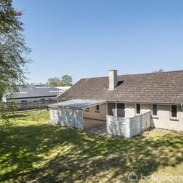 Et ældre hus står i en grøn have med en terrasse ved siden af. Huset har et rødt tegltag og en skorsten. Det er omgivet af træer og et hegn i baggrunden.
