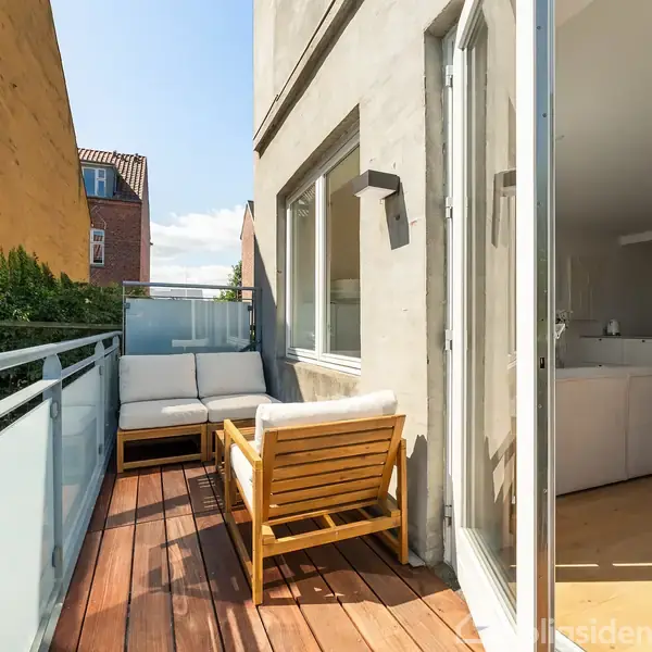 En balkon med trædæk har en sofa og lænestol opstillet mod glasrækværket. Balkonen er ved siden af en murstensbygning og åbner ind til en lys stue.