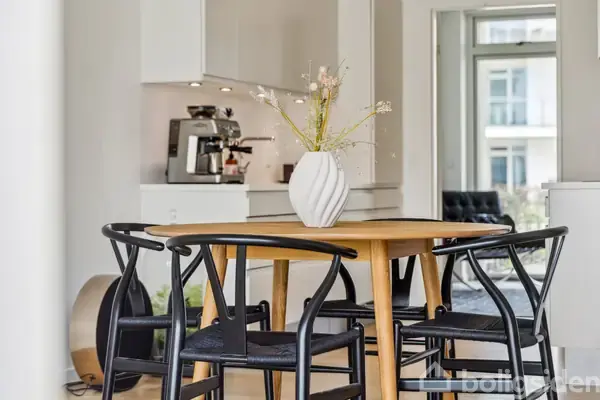 Vase med blomster står på rundt træbord omgivet af sorte stole. I baggrunden ses et moderne køkken med en kaffemaskine og hvide skabe. Lysindfald fra store vinduer skaber en åben atmosfære.
