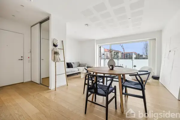 Et rundt træbord med sorte stole omkring. Bordet står i en lys stue med trægulv. I baggrunden ses en sofa nær en stor glasdør.