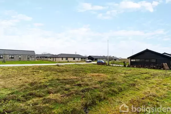 Græsbevokset tomt ligger ubenyttet i et boligområde; til venstre ses moderne huse med flade tage og i baggrunden en bil parkeret langs vejen under en klar himmel.