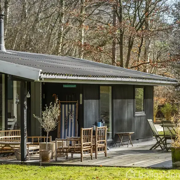 Et sort sommerhus med store vinduer og en brændeovn står i en skovklædt have. Træmøbler og planter er placeret på træterrassen, omgivet af efterårsløv og træer.