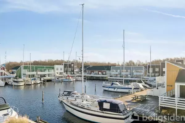 En sejlbåd ligger fortøjet ved en træbro i en marina. Omkringliggende kontekst inkluderer flere både og moderne bygninger langs kajen under en klar, blå himmel.