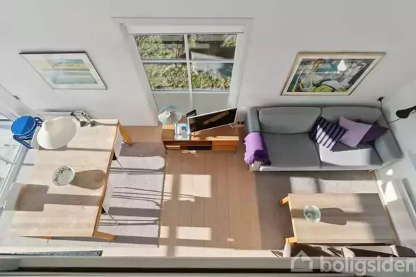 Stue set fra oven, med en grå sofa og sofabord til højre, spisebord til venstre. Vindue med udsigt til have, to malerier på væggen.