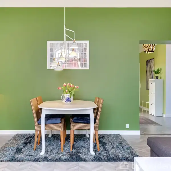 Et spisebord står med fire stole rundt om det; en vase med blomster er på bordet. Rummet har en grøn væg og en hængende lampe over bordet.