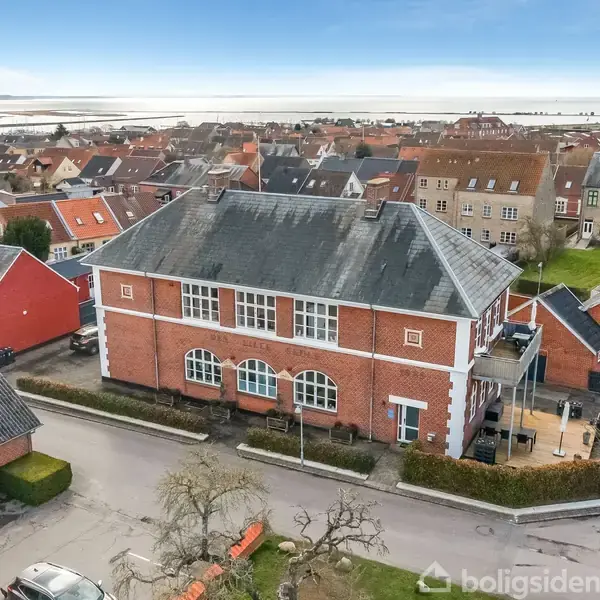 En stor, rød murstensbygning med hvide detaljer står ved en vej i et boligområde med huse omkring. Boldhavet ses i baggrunden under en klar blå himmel.