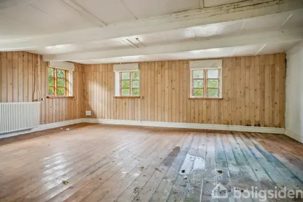 Et tomt rum med trævægge og trægulv, der har synlige pletter. Rummet har tre små vinduer, og et radiator står under det venstre vindue. Ingen tekst er synlig.