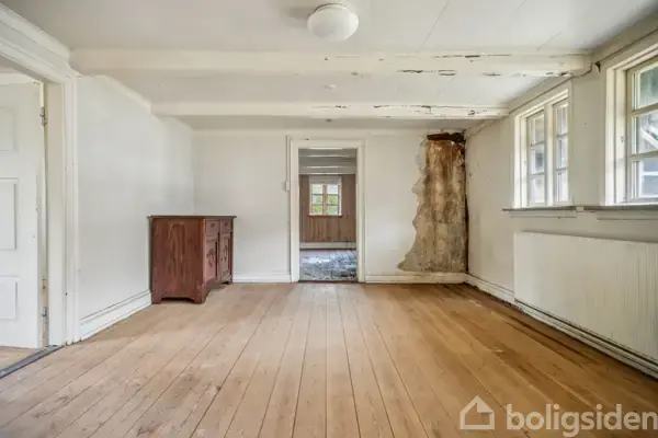 Et tomt rum har et rustikt skab langs den ene væg, et hullet område i væggen, og træplankegulv. Store vinduer bringer lys ind, mens en åben dør leder til et tilstødende rum.