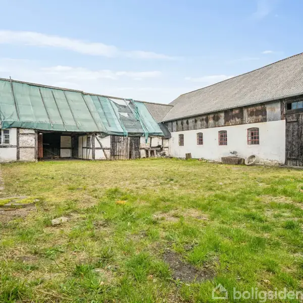 Gårdsplads med græs bevokset gårdsplads omgivet af gamle landbrugsbygninger med træ- og murstensfacade. Bygningerne har store træporte og et grønt halvtagelement. Himmel med lette skyer ses i baggrunden.