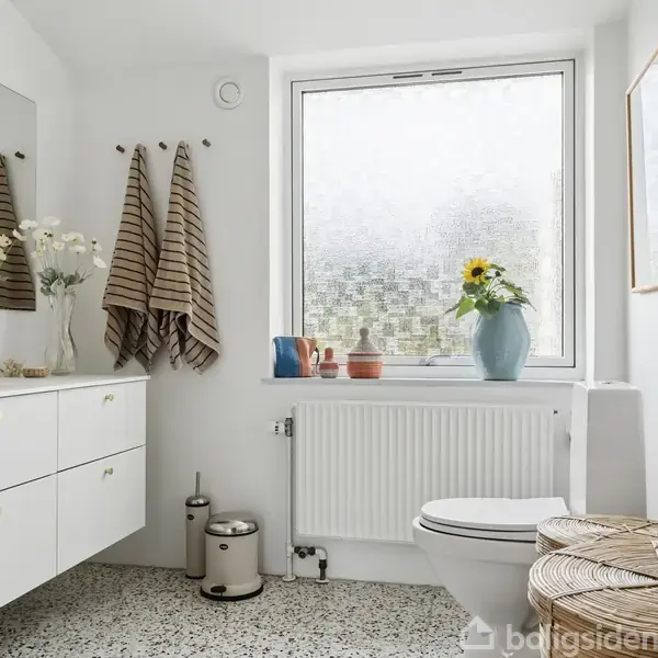 Et lyst badeværelse med hvidt skab og spejl ved siden af en vindueskarm. Håndklæder hænger på væggen, blomster står i en vase, og en vasketøjskurv er ved toilettet.
