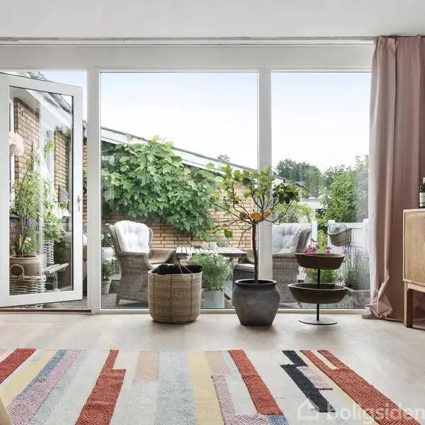 Stue med moderne møbler, herunder en brun puf og glasbord. Store vinduer med lyse gardiner giver udsigt til en have. Et lille skab står ved væggen, og planter pynter rummet.