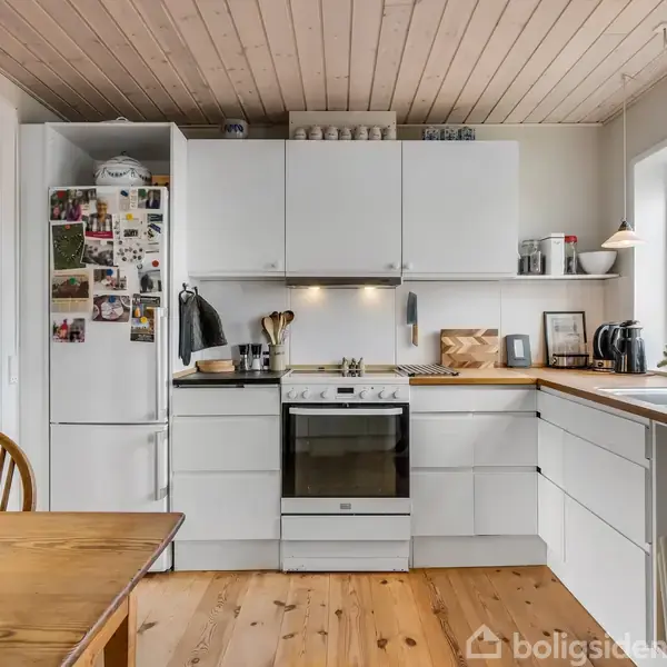 Et køkken med hvidt køkkenudstyr og trækbord, dekoreret med billeder og magneter på køleskabet. Sollys strømmer ind gennem vinduerne, og der hænger jakker ved døren.