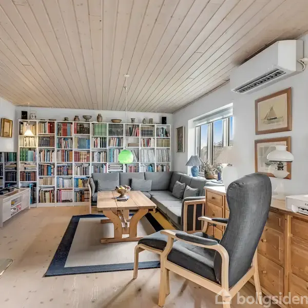 Reol rummer bøger; sofagruppe omkring sofabord; hyggelig brændeovn; skrivebord med printer og kontorstol; vægkunst med skibsbilleder; lys indretning med vindue, aircondition og trægulv.