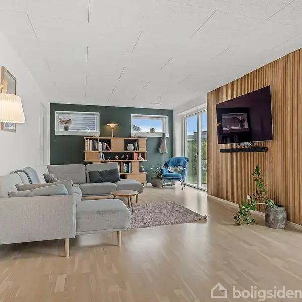 Grå sofa og lænestol står i moderne opholdsstue med træpanelvæg med monteret fladskærm. Store vinduer viser terrasse med havemøbler udenfor. Grøn plante står ved væggen.