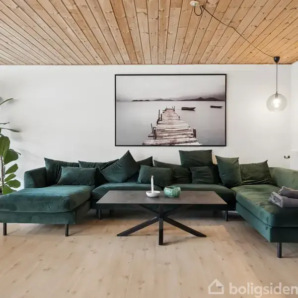 Stor, mørkegrøn sofa med puder står i en moderne stue. På væggen hænger et sort-hvidt billede af en bro. Et elegant spisebord og en høj plante giver indretning dybde.