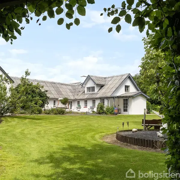 Hus beliggende på en stor, velplejet græsplæne omgivet af træer og buske. Bygningen har en hvid facade og grå tag, med blå himmel i baggrunden.