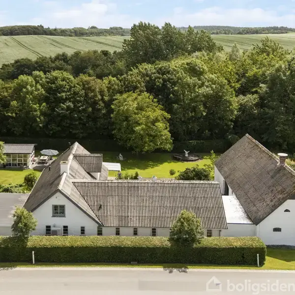 En hvid, stråtækt bygning står stille i en grøn landlig setting, omgivet af tæt skov og marker. En vej løber foran bygningen, adskilt af en hæk.