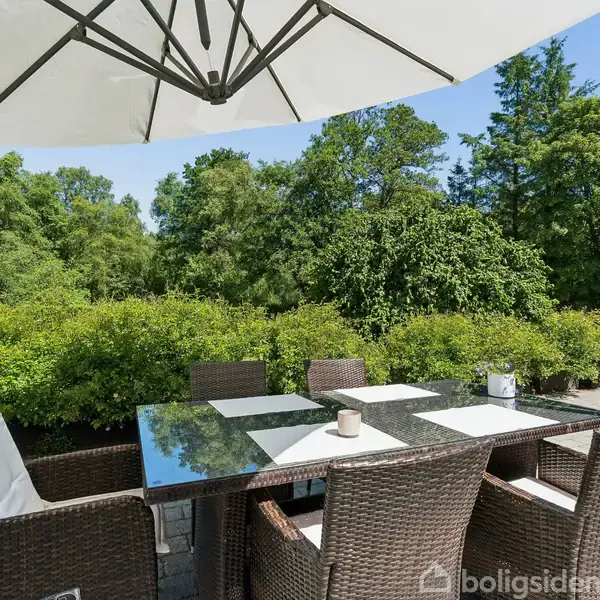 Rundt havebord med glasplade, omgivet af flettede stole, står under en stor parasol. Stedet er en terrasse med udsigt til frodig, grøn beplantning.