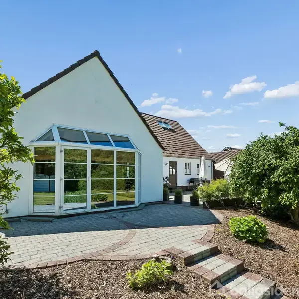 En hvid, moderne villa med store vinduespartier står i en velplejet have. Træer og buske omgiver huset, mens solen skinner fra en blå himmel med spredte skyer.