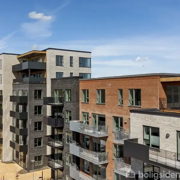 Moderne lejlighedskompleks står stille, omgivet af åbne områder under en blå himmel. Bygningerne består af flere etager med altaner, facader i grå, brune og sorte nuancer.