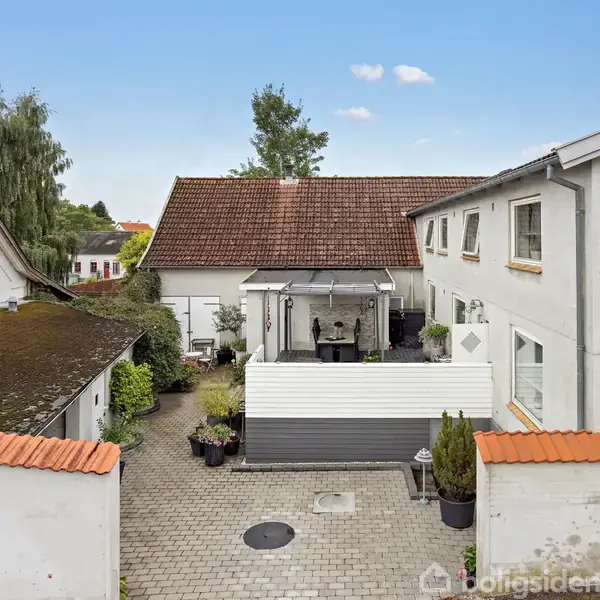 Et gårdhaveområde med brostensbelægning er omgivet af hvide huse med røde tegltage. En overdækket terrasse har havemøbler og planter. Spredte krukker pryder det velholdte område.