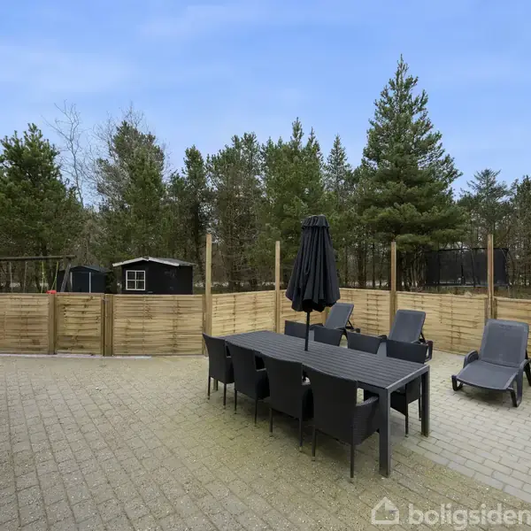 Terrassemøbler står på en brostensbelagt terrasse med et langt bord, otte stole og to liggestole. Parasol og træhegn omgiver området, med en baggrund af høje, grønne træer.