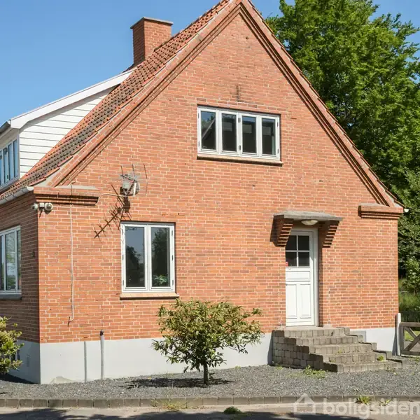 Rødt murstenshus med gavltag står stille i en forstadshave. Trappetrin fører op til hoveddøren, omgivet af buske. Store vinduer ses på facaden mod lyseblå himmel.