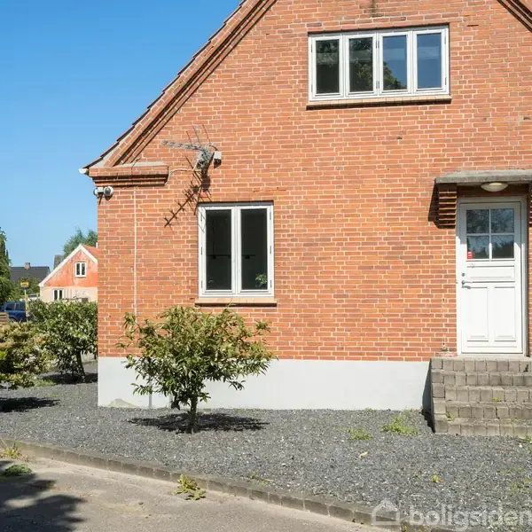 Et rødt murstenshus med en hvid dør og to vinduer står i solen. Foran er små buske og en grusbelagt have. Huset er omgivet af en vej og nabohuse.